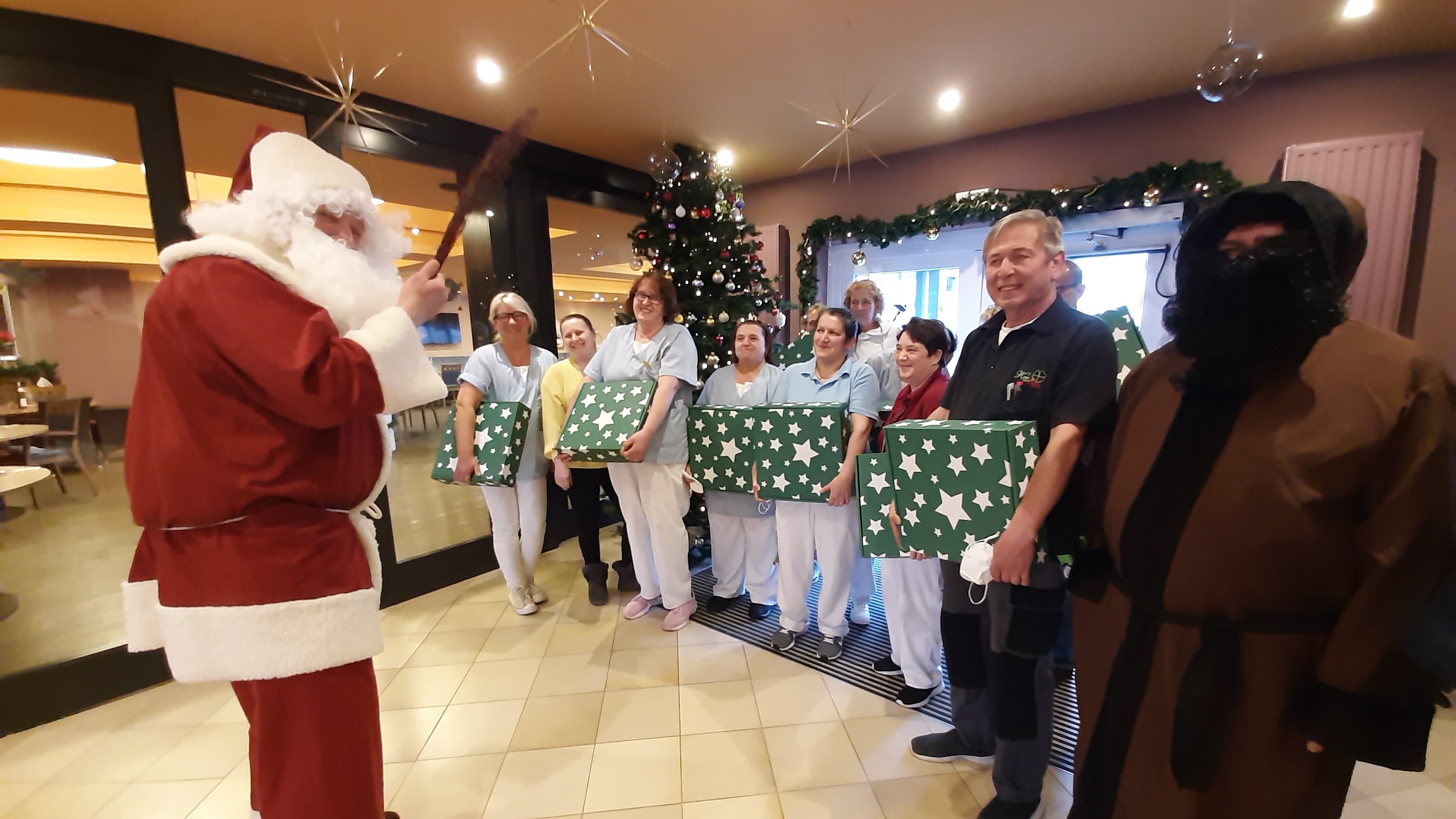 Ein als Weihnachtsmann verkleideter Mann steht vor einer Gruppe von Pflegekräften in weißer Kleidung, die Geschenke mit grünem Papier und goldenen Schleifen halten. Im Hintergrund steht ein festlich geschmückter Weihnachtsbaum. Die Szene vermittelt festli